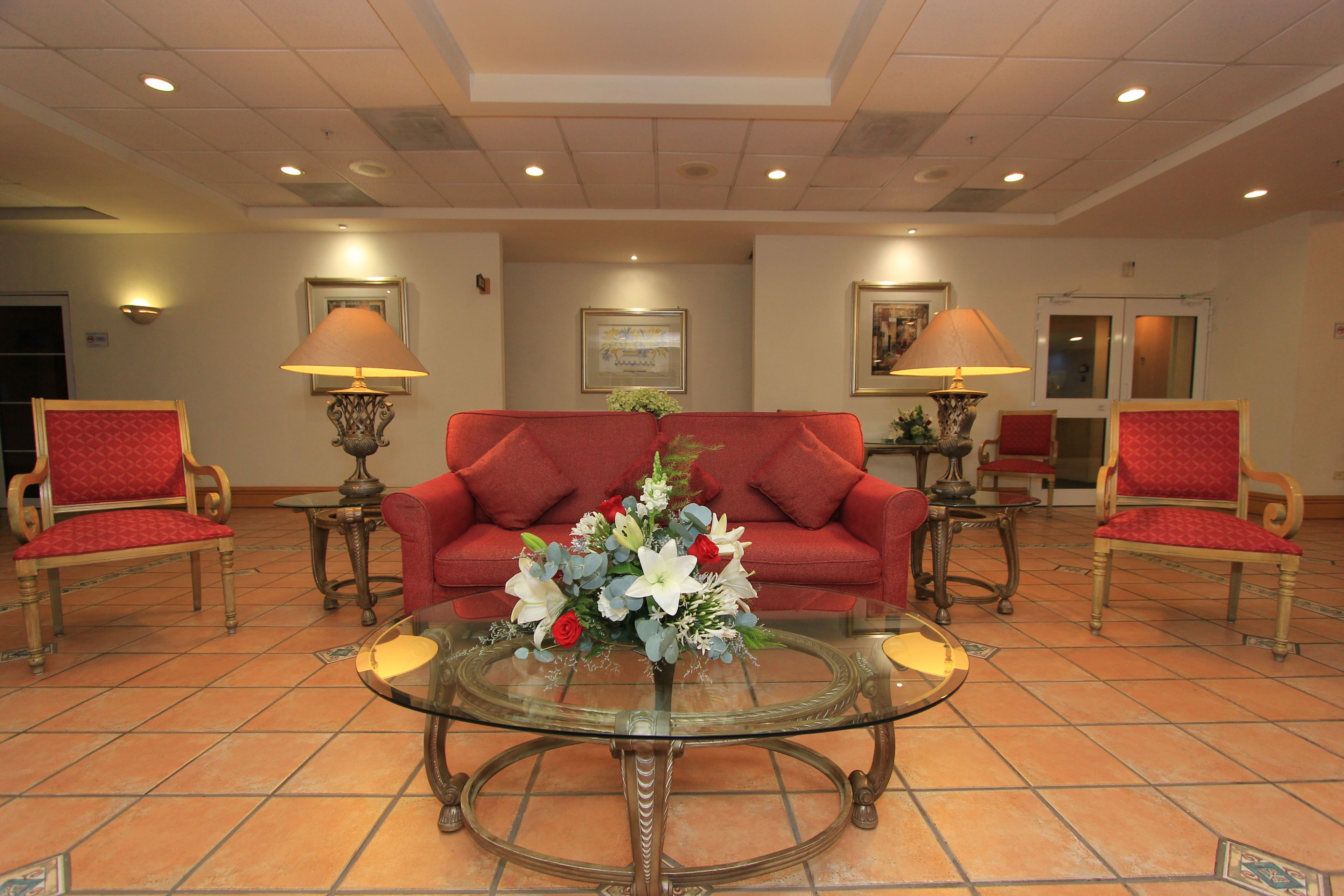 A lobby with a red sofa, two armchairs, a glass coffee table with a floral centerpiece, and two floor lamps.