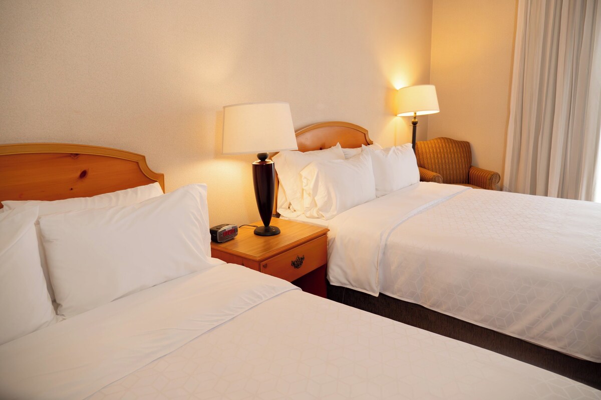 A hotel room with two beds, a nightstand, a lamp, and a chair.