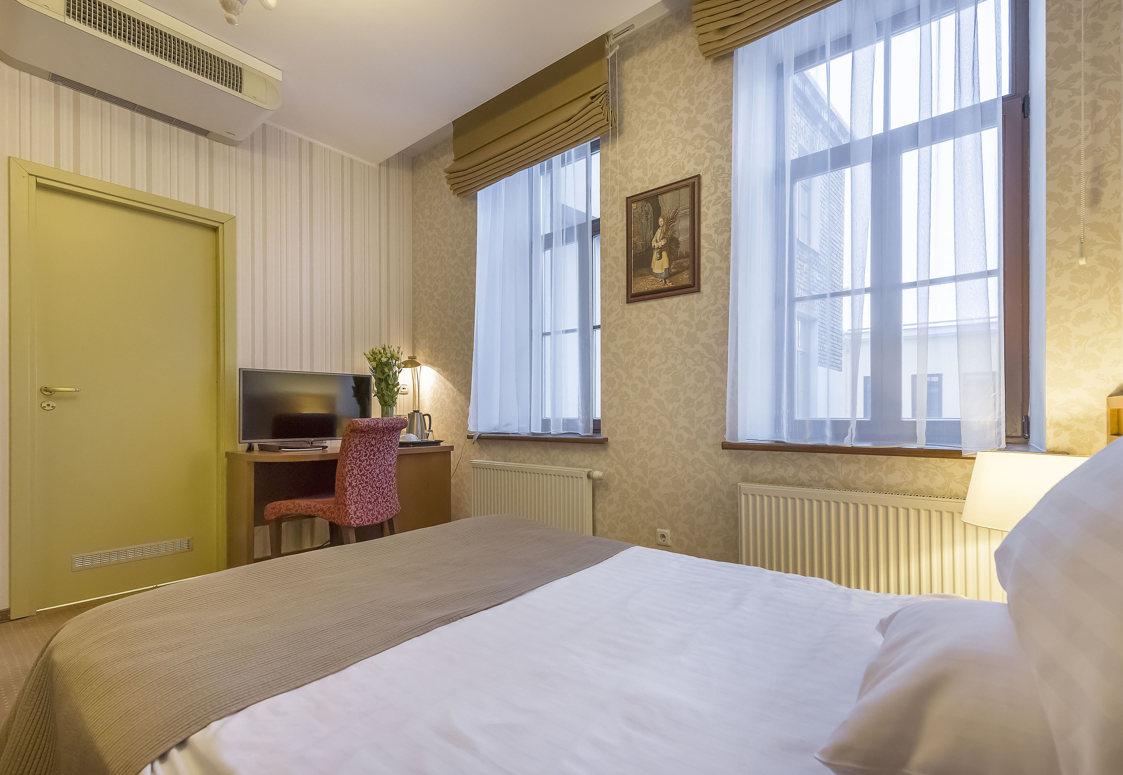 Hotellihuone, jossa on sänky, työpöytä tietokoneella, tuoli ja ikkuna, jossa on verhot.