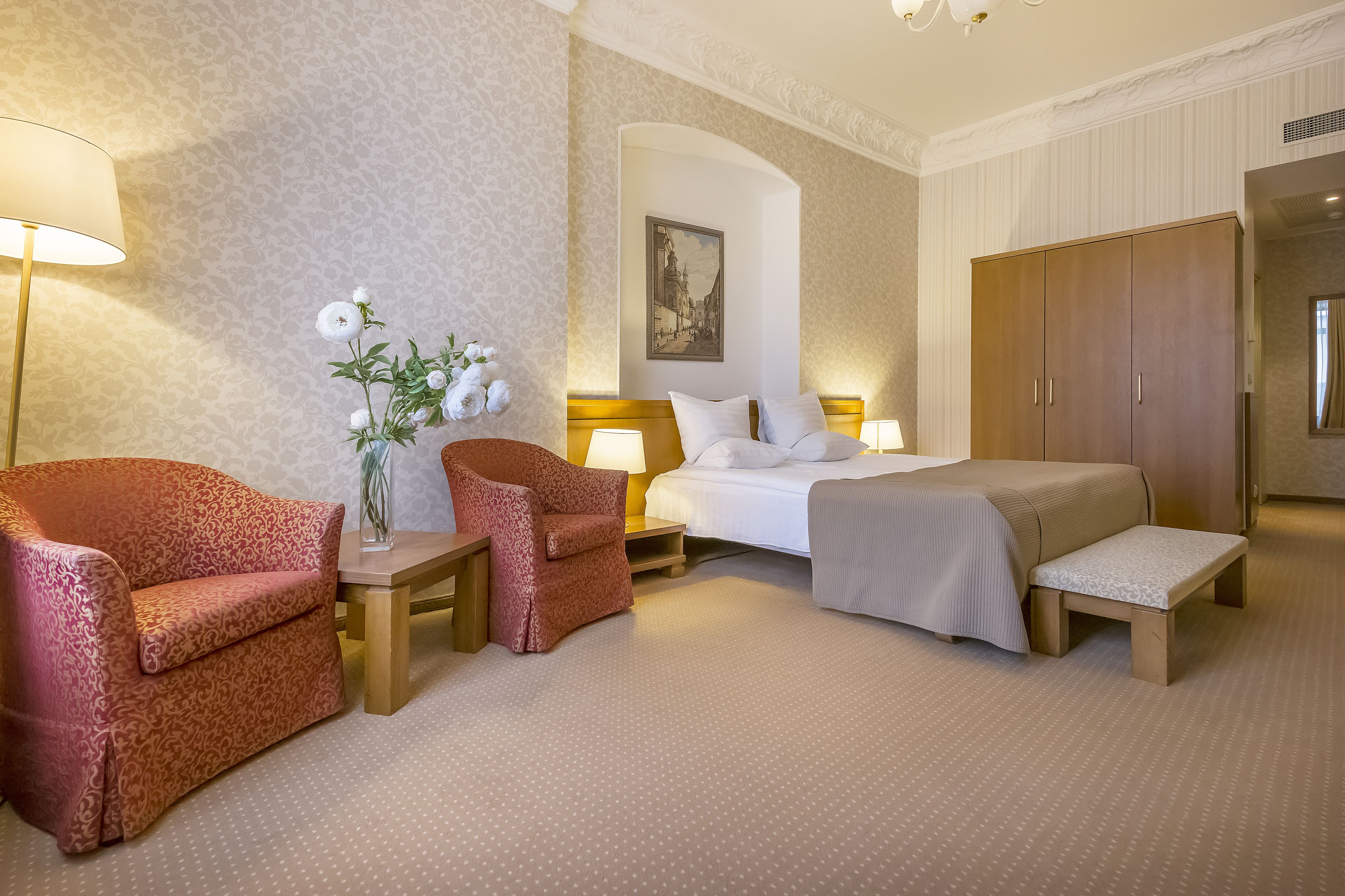 A hotel room with a bed, two armchairs, a small table with a vase of flowers, a lamp, and a wardrobe.