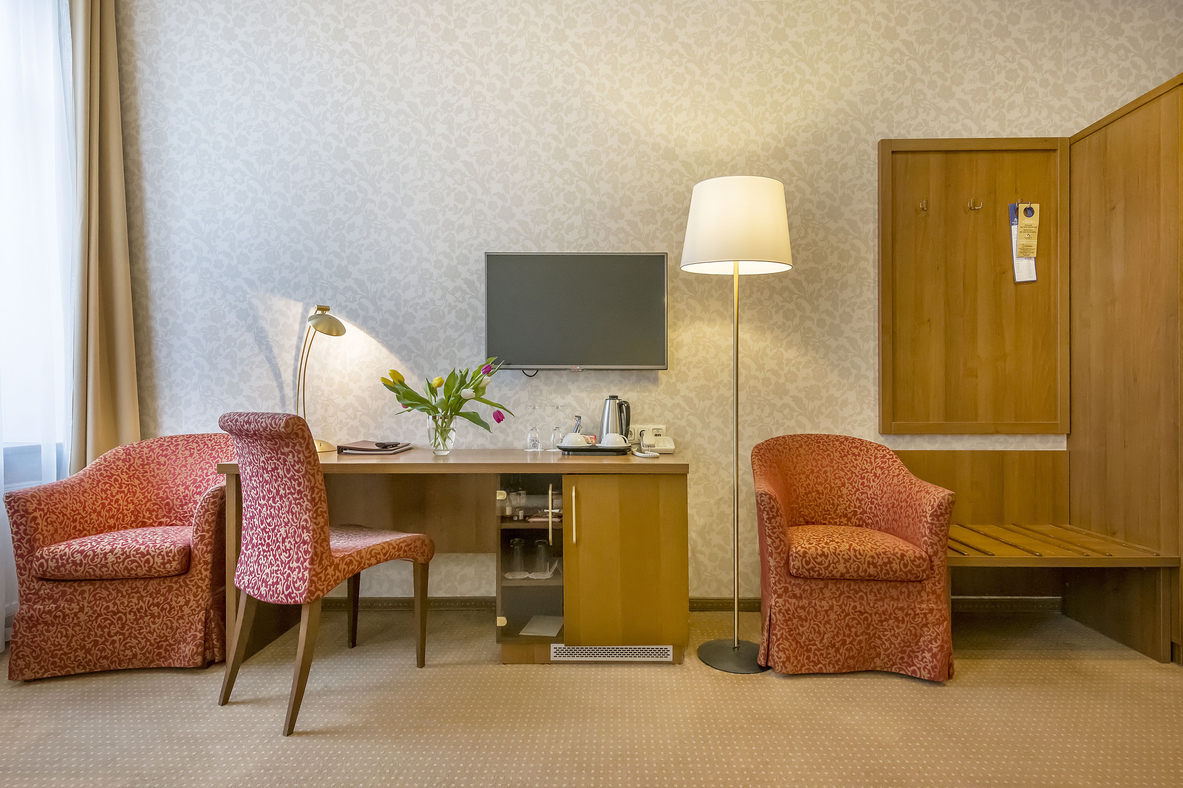 Habitación de hotel con escritorio, dos sillones estampados, lámpara, televisor y armario.