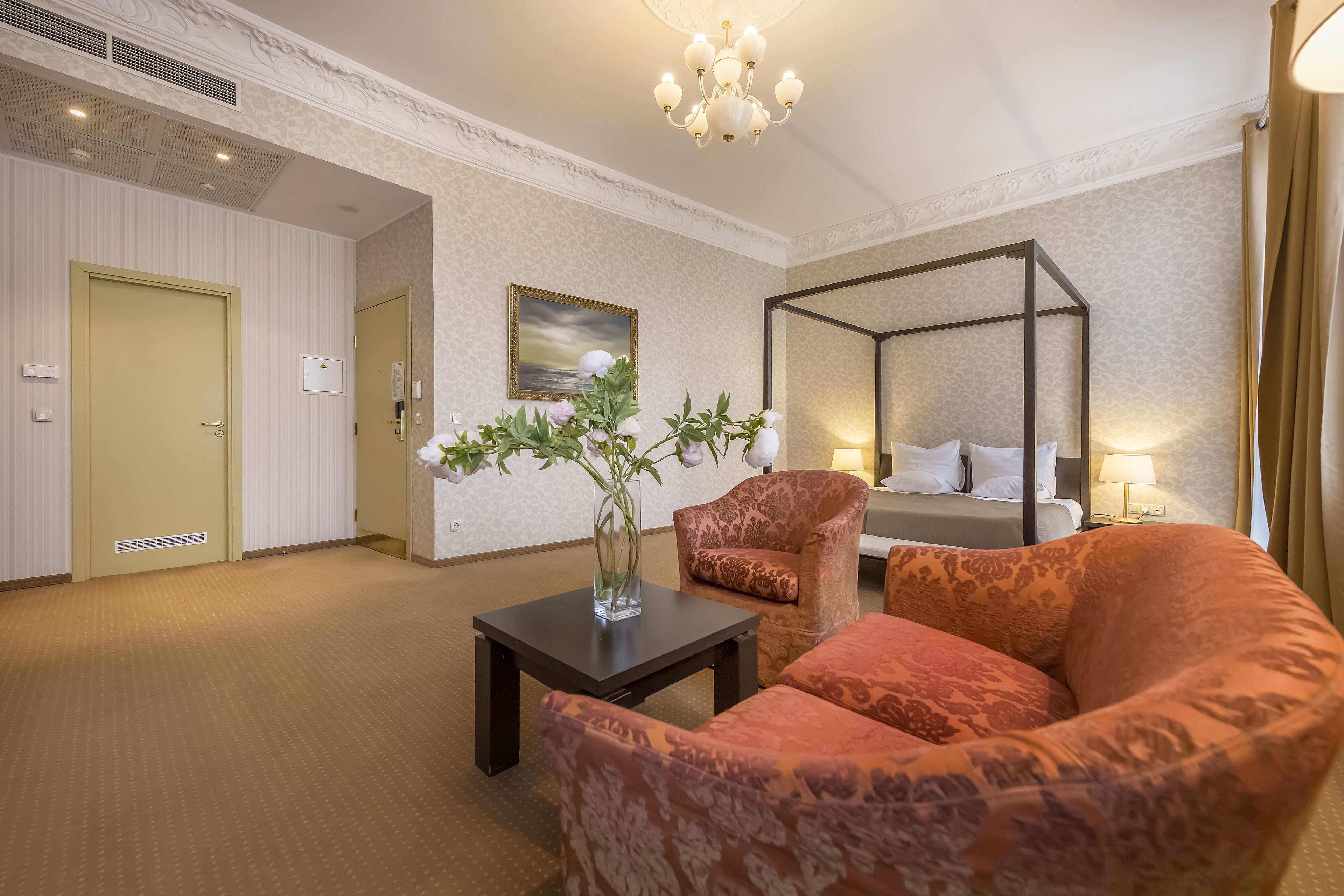 Habitación de hotel con cama con dosel, zona de estar con sillón estampado, una mesita con un florero y un cuadro en la pared.