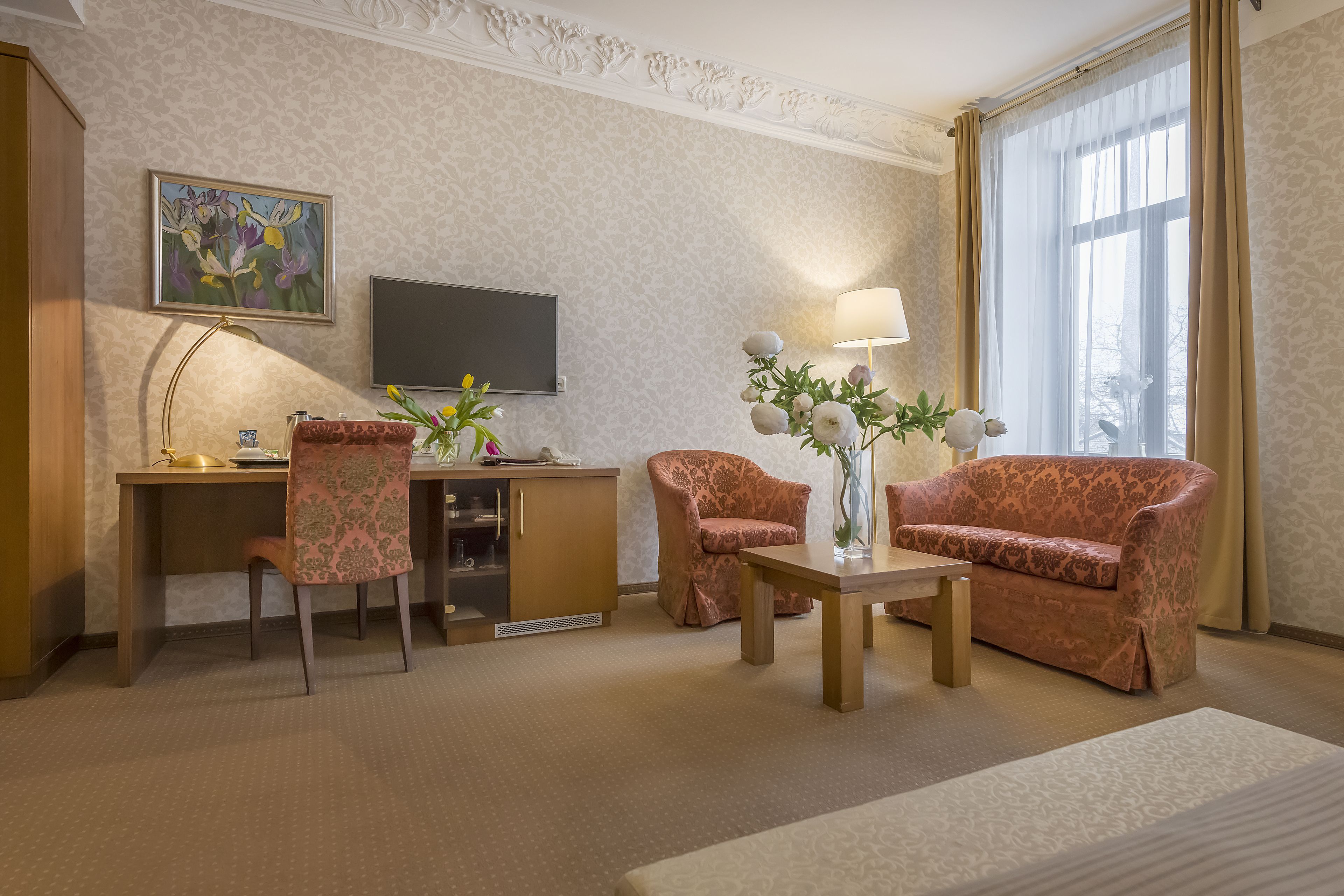 Une chambre d’hôtel équipée d’un bureau, de deux fauteuils à motifs, d’un canapé, d’une télévision et d’une fenêtre avec des rideaux.