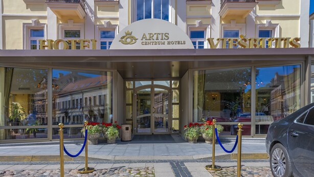 The entrance of the Artis Centrum Hotels with a clear glass facade, a welcoming archway, and a blue rope barrier.