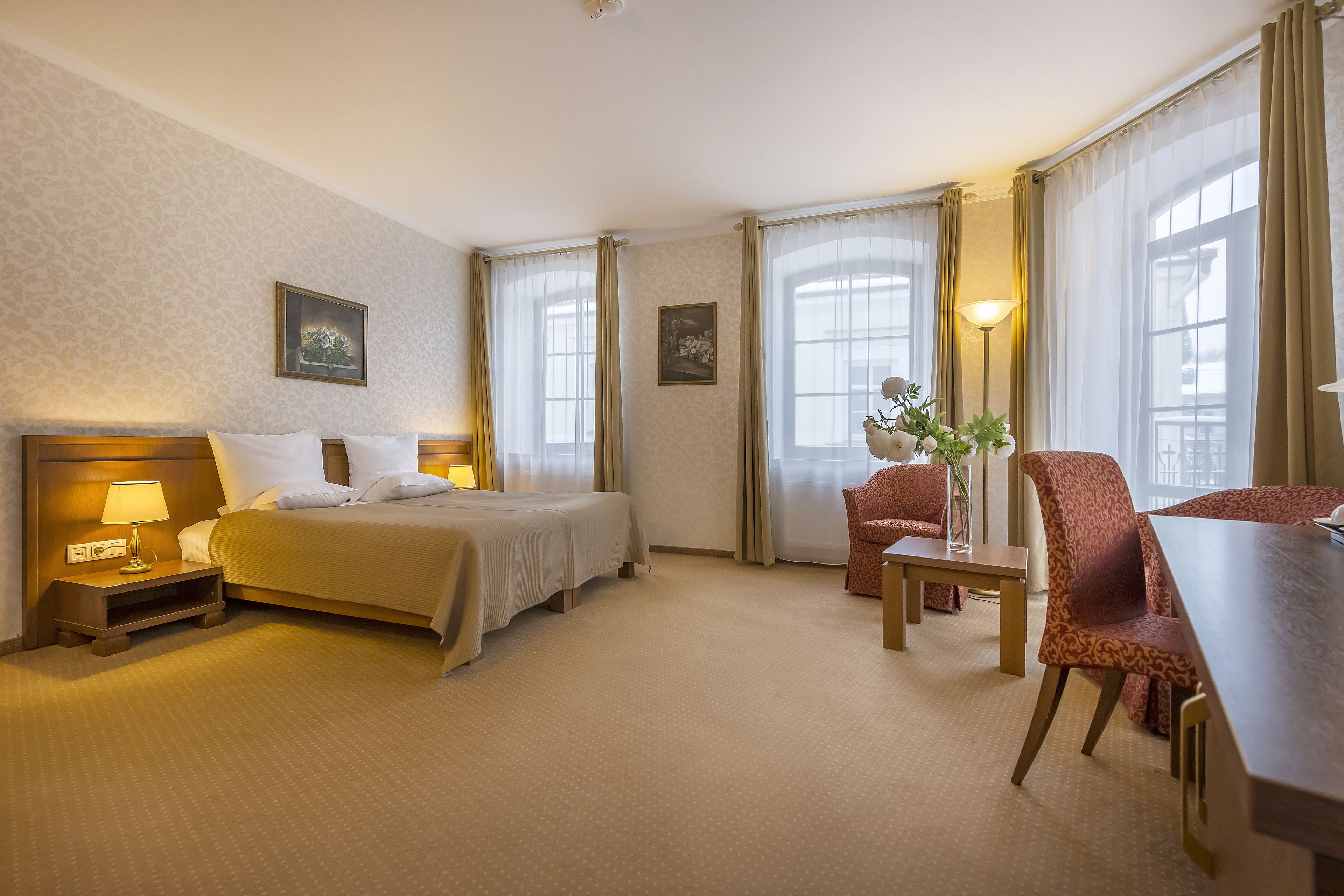 Ein Hotelzimmer mit Doppelbett, Schreibtisch mit Stuhl, einem kleinen Tisch mit Vase und zwei mit Vorhängen versehenen Fenstern.