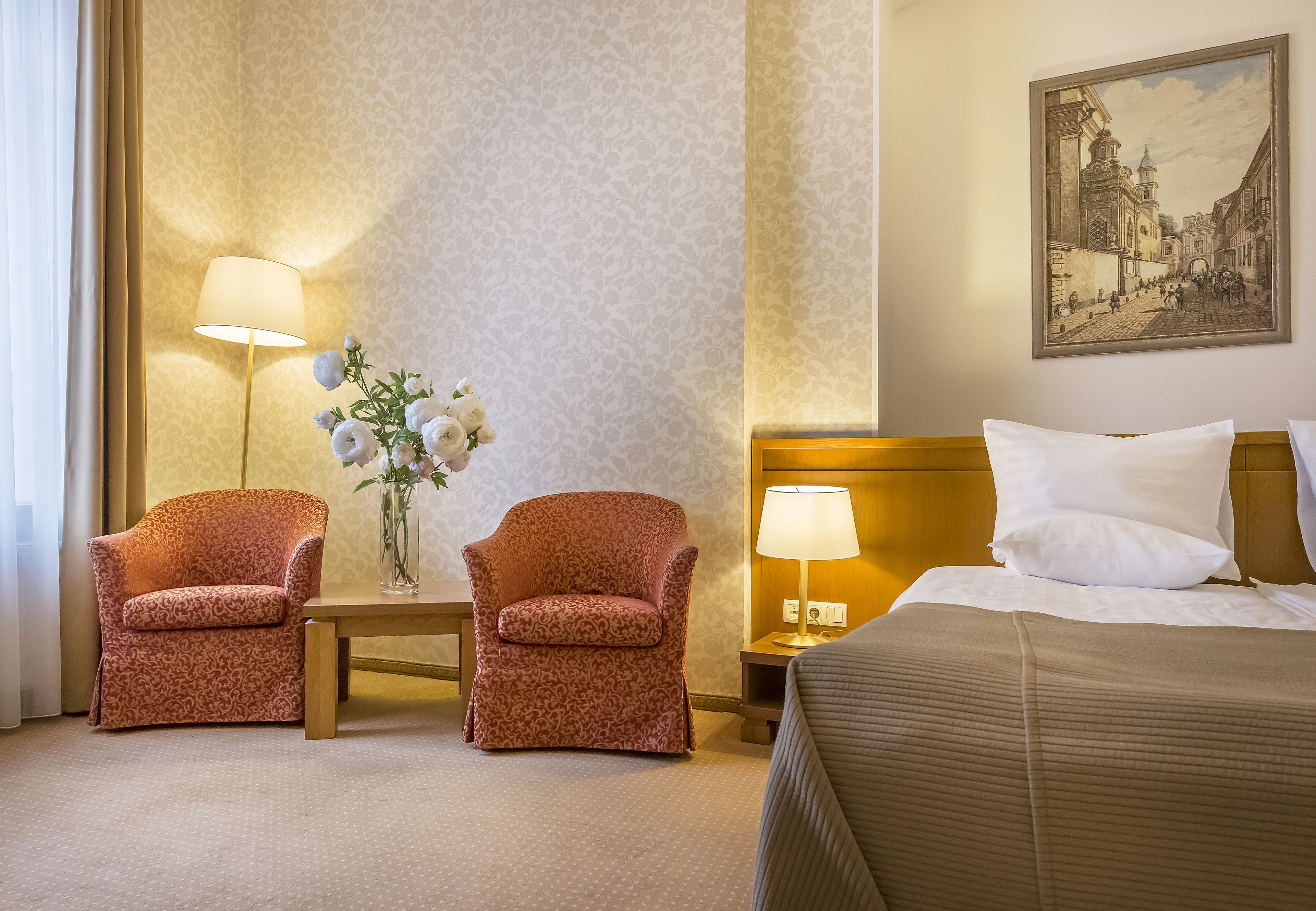 A hotel room with two armchairs, a small table with a vase of flowers, a lamp, and a bed with a headboard.