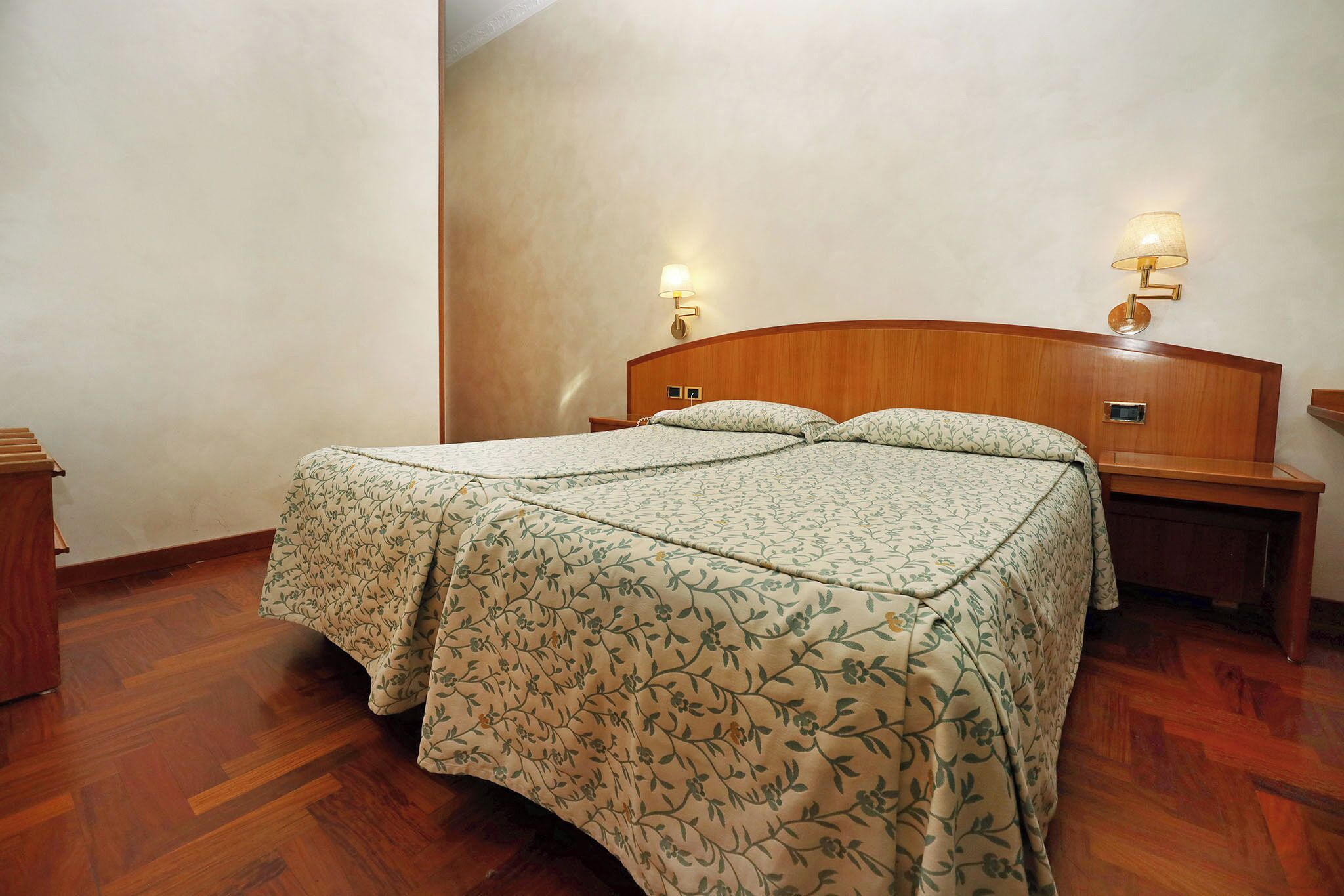A hotel room with a wooden headboard, two bedside lamps, a bed with floral bedding, and a wooden nightstand.