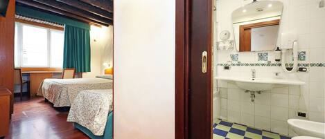 Una habitación con techo de madera, un escritorio, dos camas con cubrecamas florales, una ventana con cortina verde y un baño con piso a cuadros, azulejos blancos y un lavamanos.