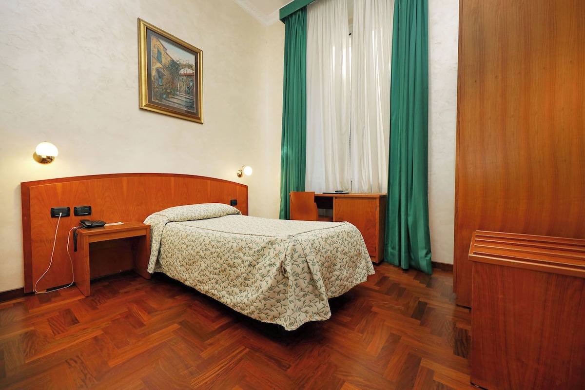 A neatly arranged bedroom with a wooden headboard, a bedside table with a lamp, a bed with a floral bedspread, a painting on the wall, and a window with green curtains.