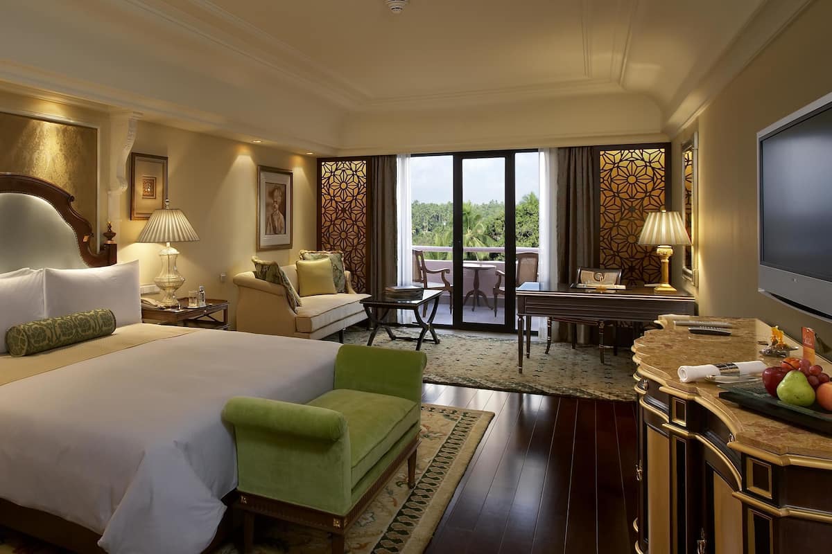 Habitación de hotel con una cama grande, un sillón verde, un escritorio con una silla y un balcón con vistas.