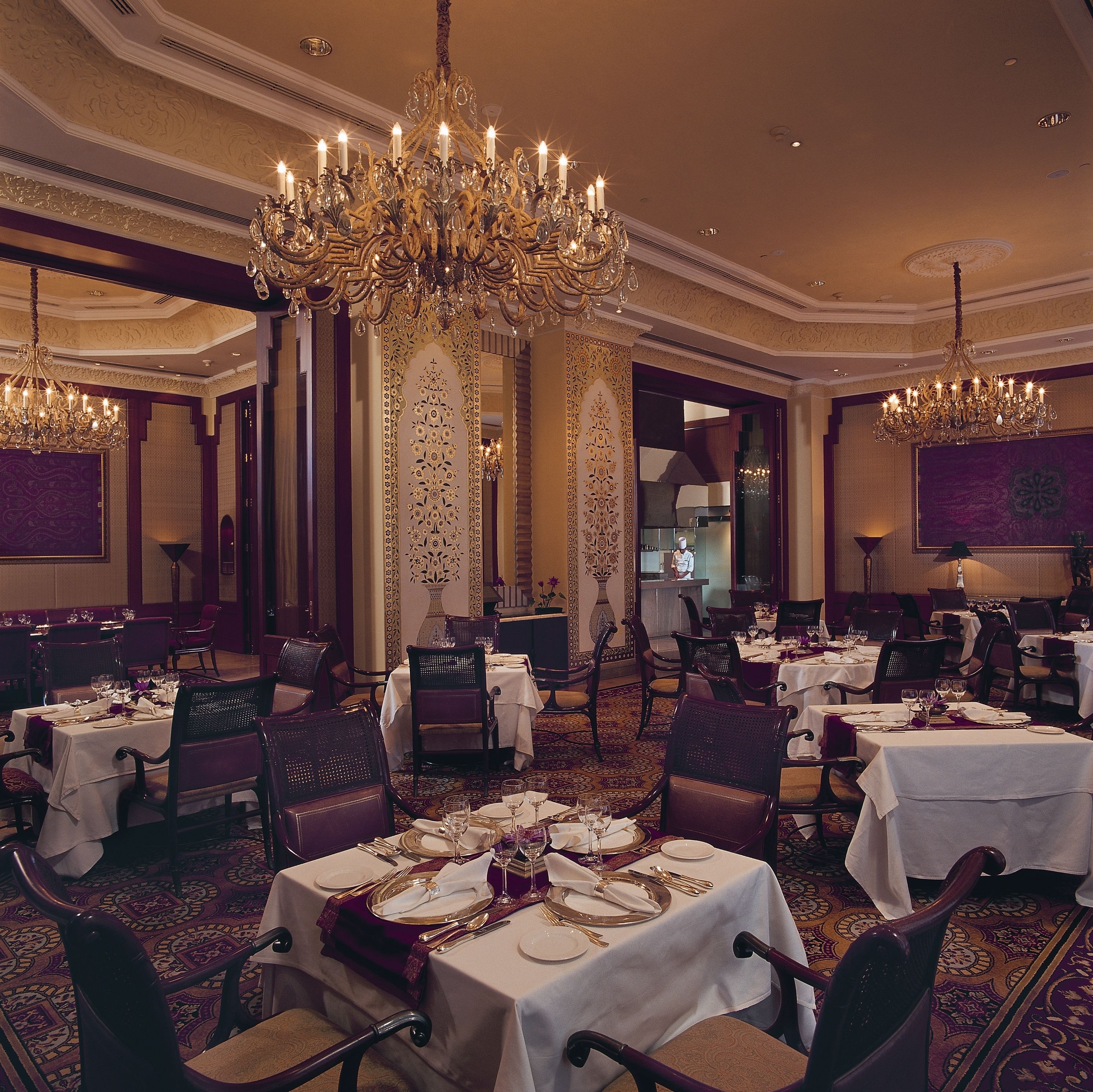 A formal dining room with multiple tables set for service, ornate chandeliers, and a classic interior design.