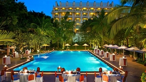 A poolside dining area with tables set for a meal, surrounded by palm trees and a large building in the background.