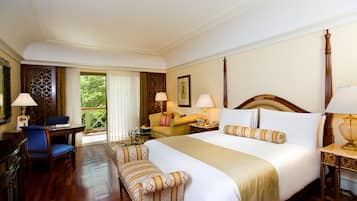 A hotel room with a large bed, a desk, a chair, a sofa, and a view of greenery outside.
