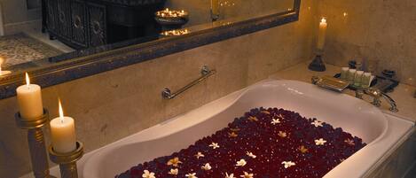 A bathroom with a bathtub filled with rose petals, a mirror, and a vanity with a sink and toiletries.