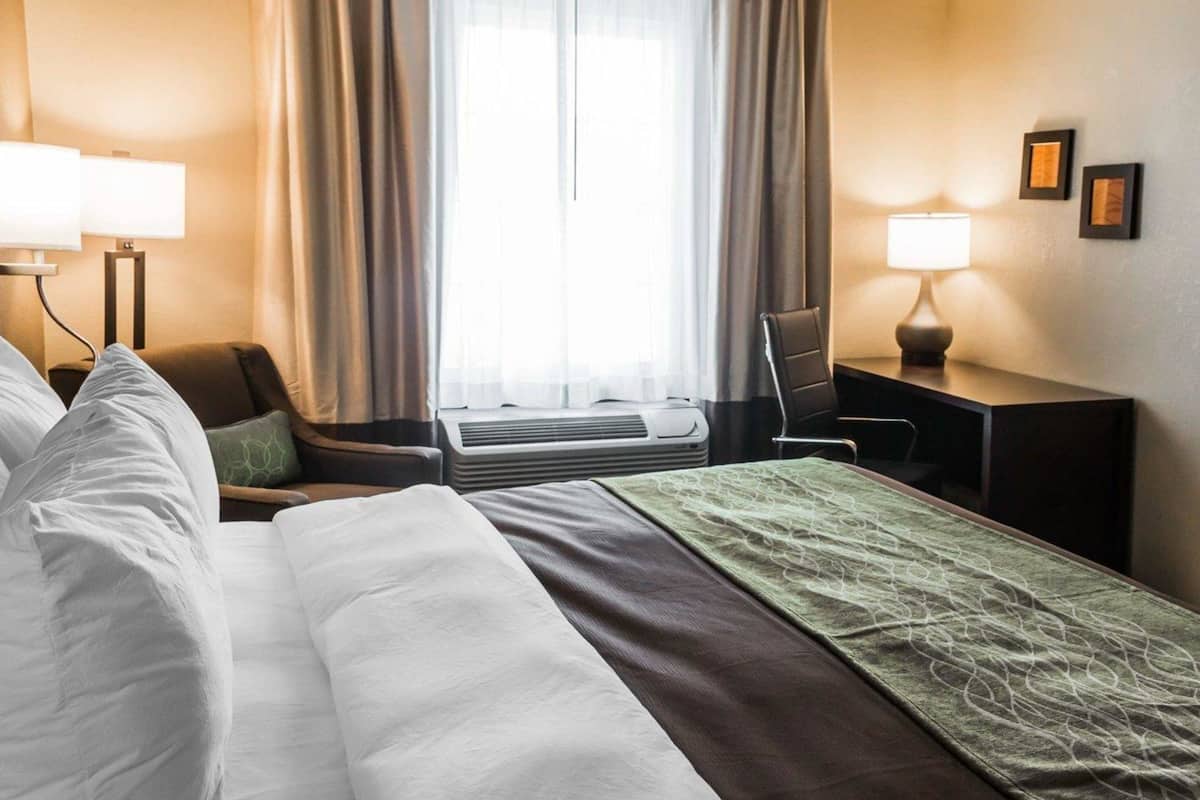 A hotel room with a bed, a desk, a chair, a lamp, and a window with curtains.
