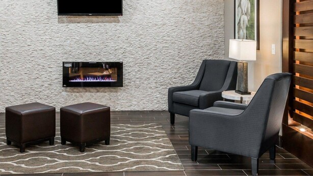 A modern living room with a stone wall, a flat-screen TV, a fireplace, two leather ottomans, and a dark grey armchair. - Comfort Inn & Suites Pharr - McAllen (Pharr)