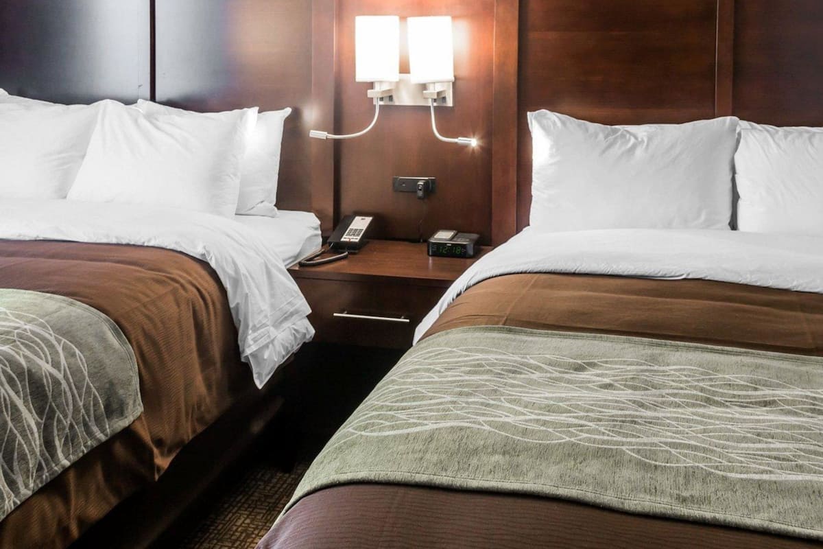 A hotel room with two beds, a nightstand with a phone and a clock, and a wall-mounted light fixture.
