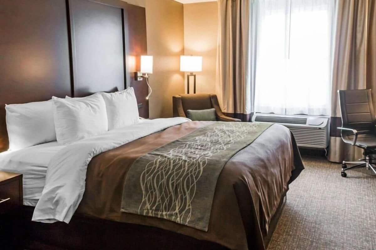 A hotel room with a large bed, two bedside lamps, a chair, an air conditioning unit, and a window with curtains.