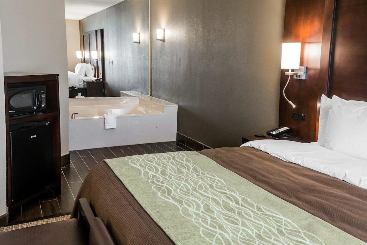 A hotel room with a large bed, a separate bathroom with a bathtub, a desk with a microwave, and a nightstand with a phone.