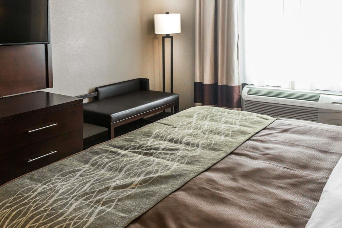 A hotel room with a bed, a desk, a chair, a lamp, and an air conditioning unit.