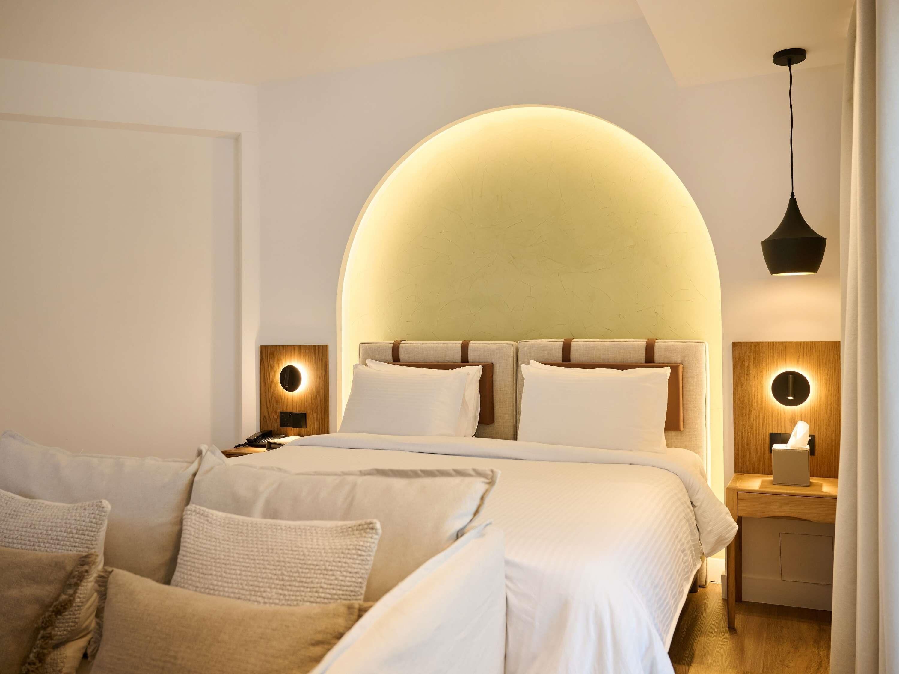 A modern hotel room with a large bed, two bedside tables, a desk, and a hanging light fixture.