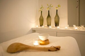 Wellnessbereich mit drei Blumenarrangements auf einem Regal, einem Holzlöffel, einer Schüssel und Kerzen auf einem weißen Handtuch.