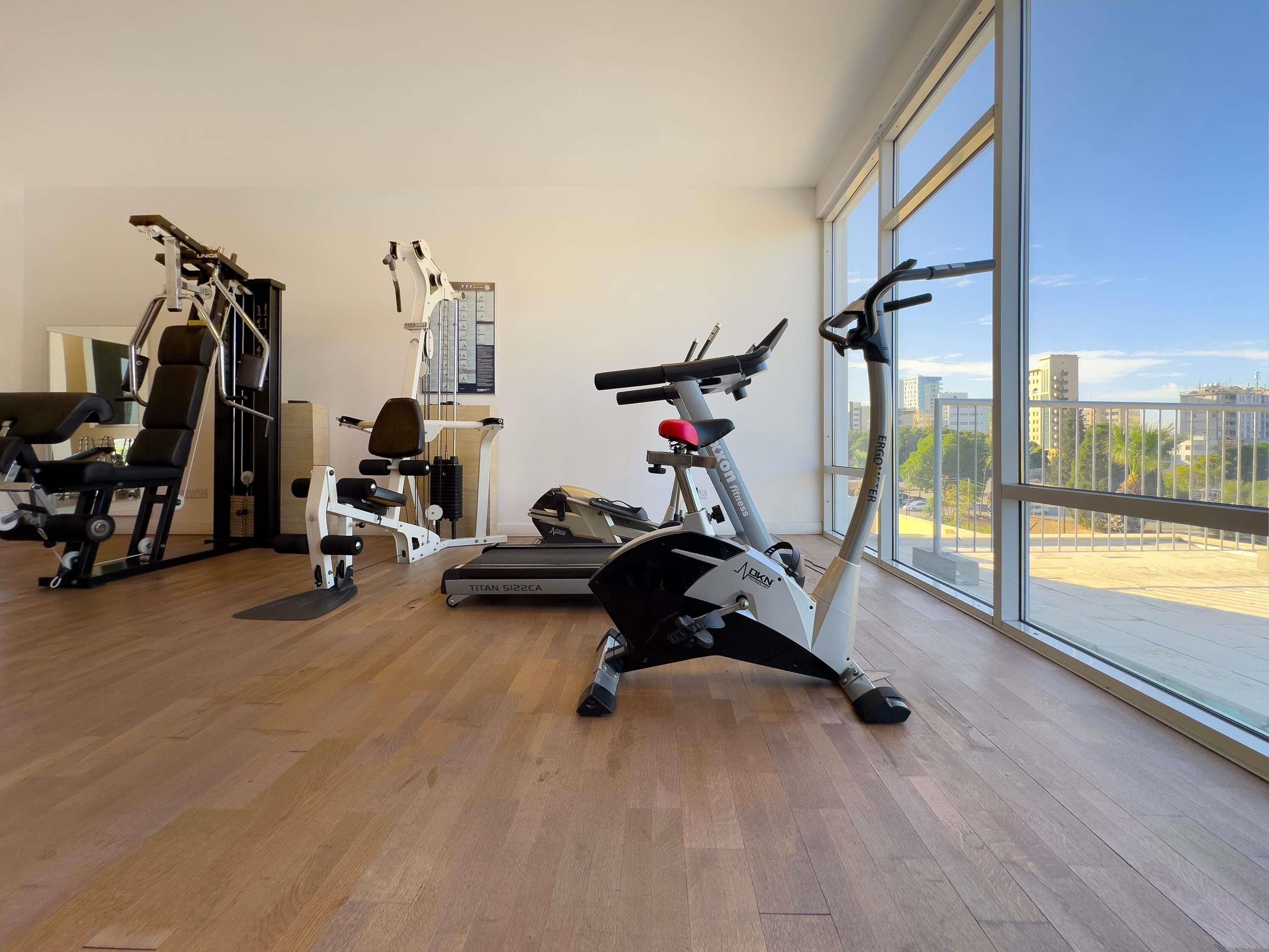 Un gimnasio con equipo de ejercicio, cinta de correr y un amplio ventanal con vistas a edificios.