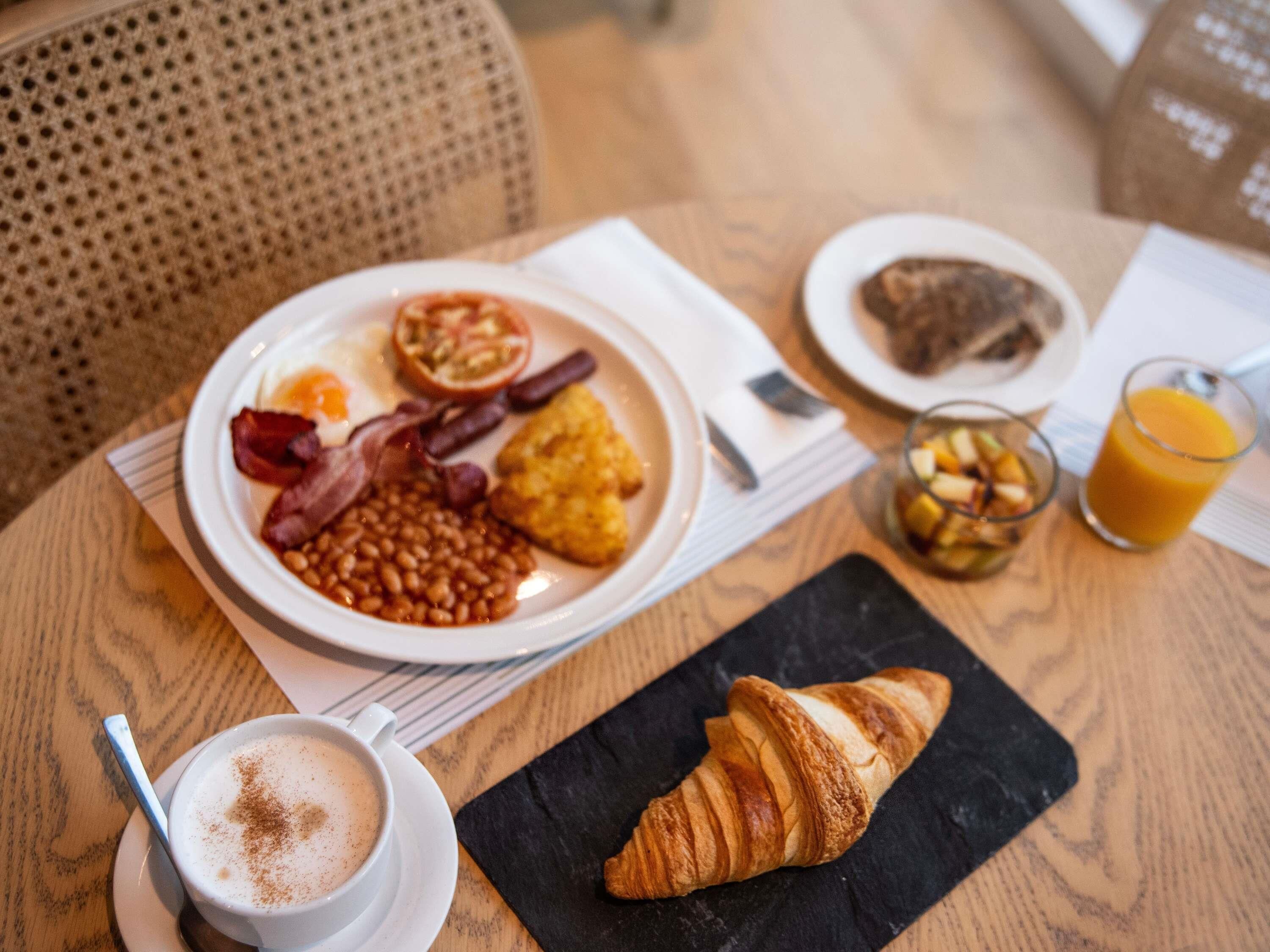 A breakfast setting with a plate of eggs, beans, bacon, and toast, a croissant, a glass of orange juice, and a cup of coffee.