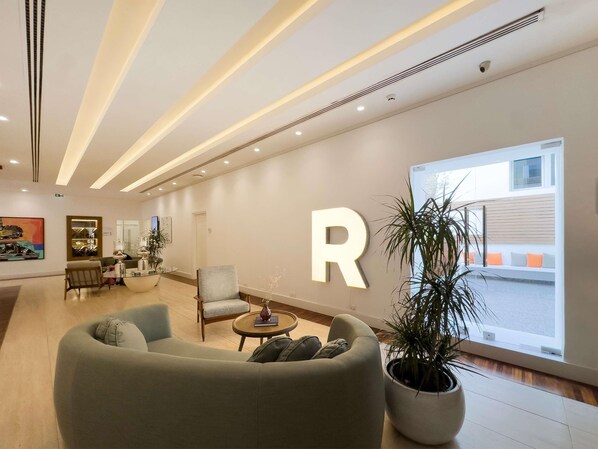 A modern lobby with a large illuminated 'R' sign, comfortable seating, a potted plant, and a view of the outside.