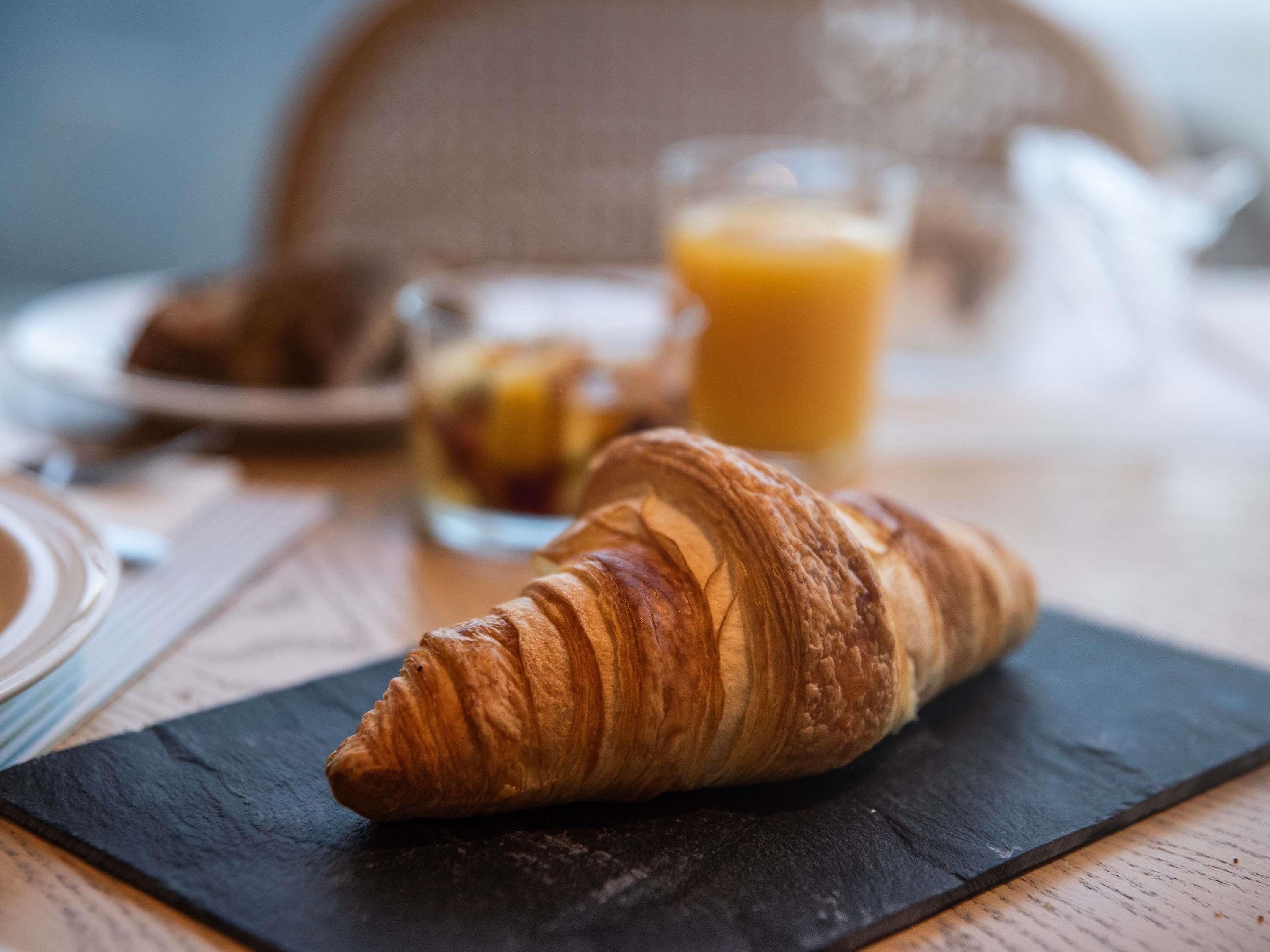 A croissant on a slate plate, a glass of orange juice, and a blurred background with a chair and a plate of food.