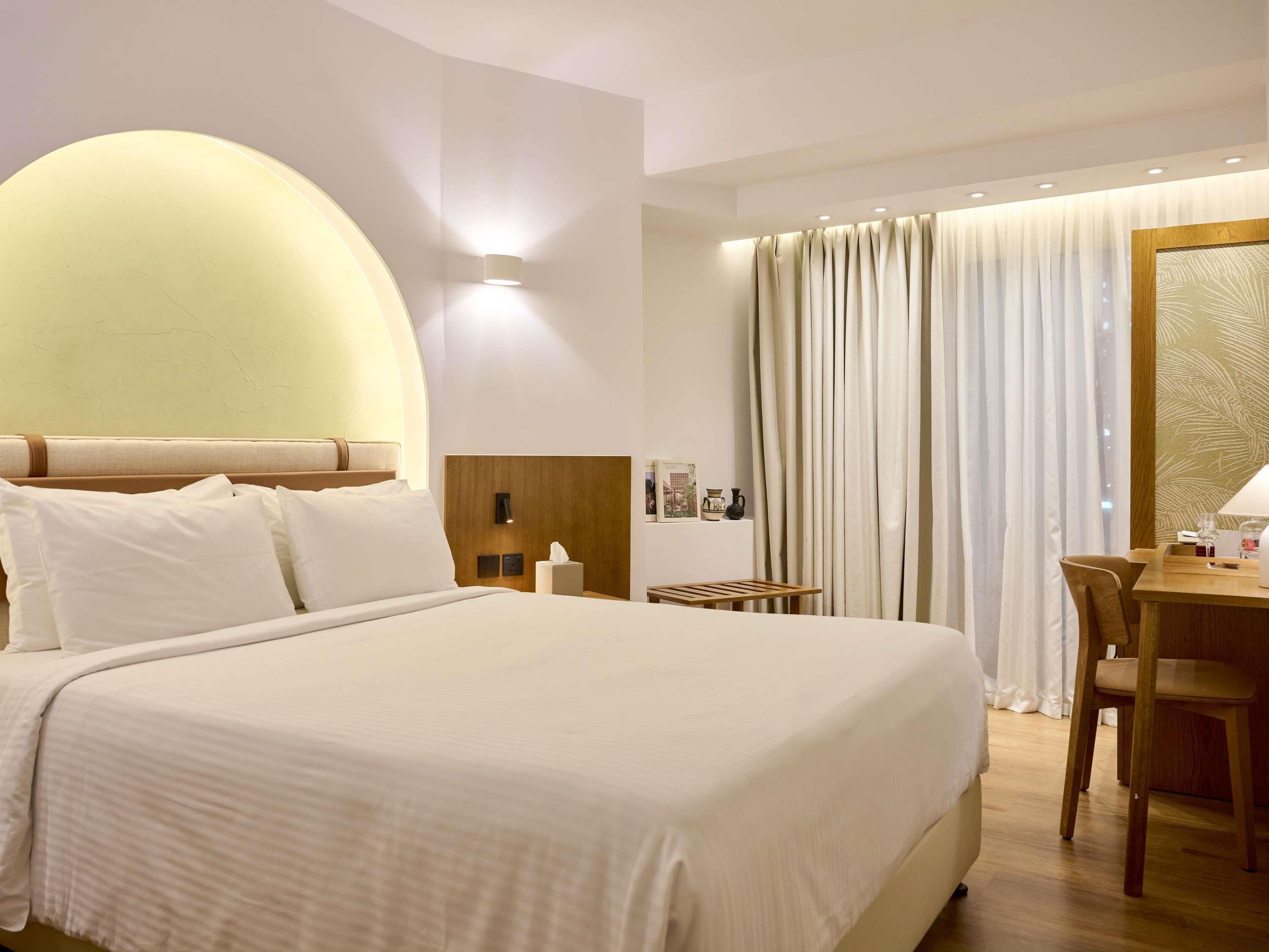 Ein Hotelzimmer mit einem großen Bett, einem Nachttisch mit Lampe, einem kleinen Tisch mit Stuhl und einem Fenster mit Vorhängen.