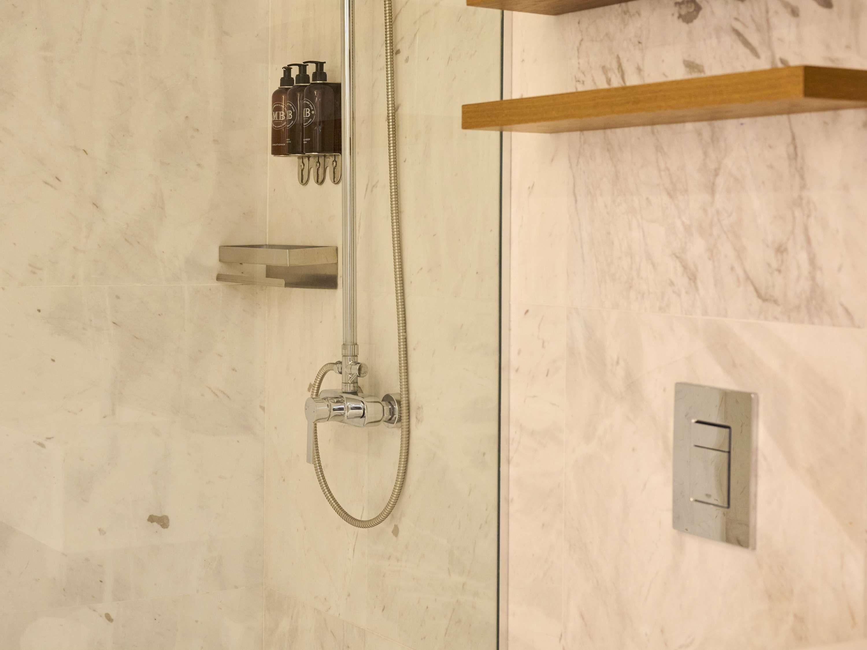 Une douche équipée d’un flexible à main, d’un panneau de commande fixé au mur et d’une étagère pour les articles de toilette.