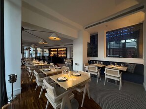 A modern restaurant with long tables, white chairs, and a bar area.