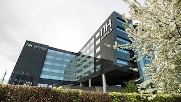 Edificio moderno de hotel con un llamativo letrero de 'NH HOTELS', amplias ventanas y un seto bien cuidado al frente.