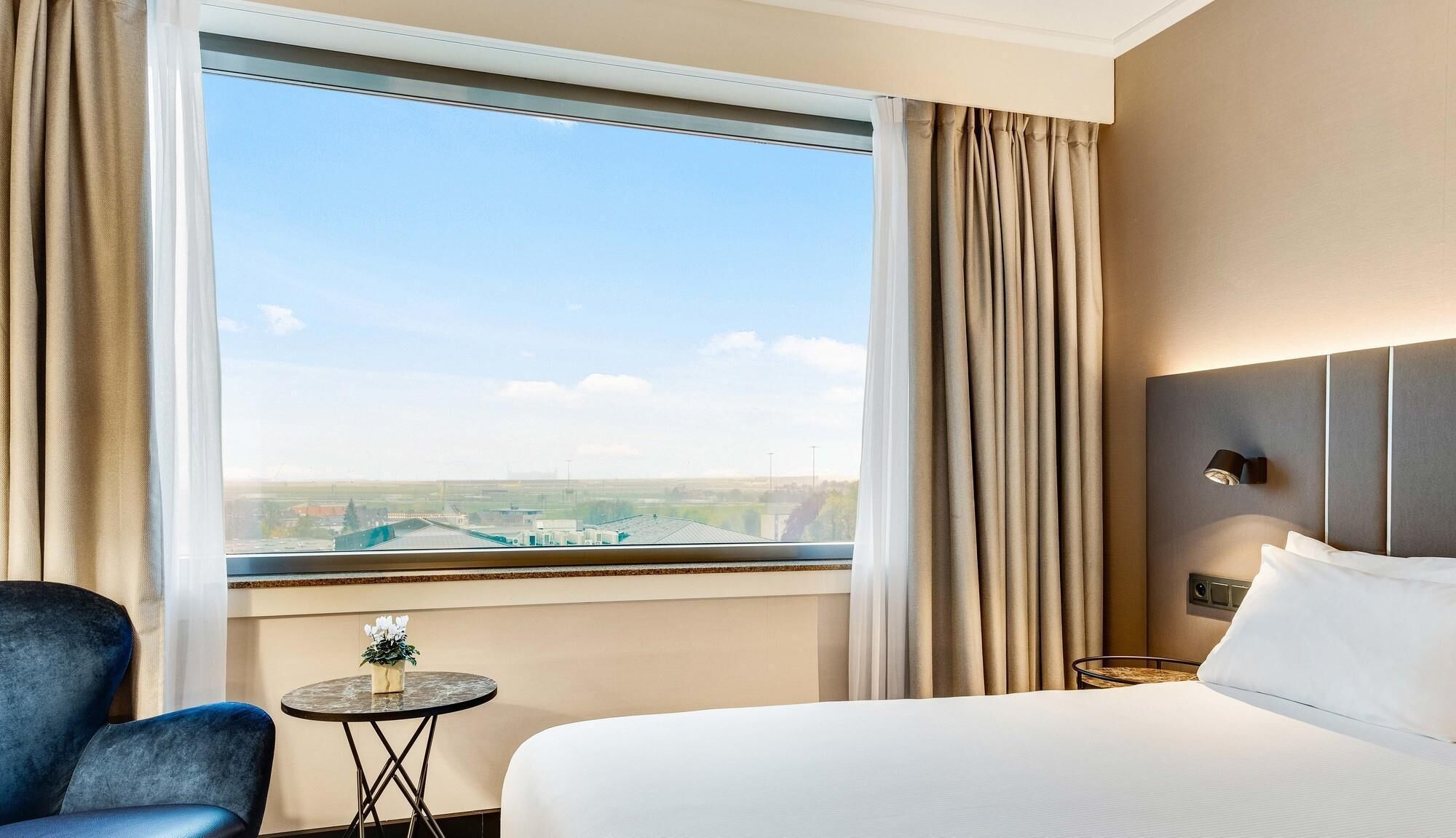 A hotel room with a bed, a chair, a small table with a plant, and a view of the cityscape through the window.