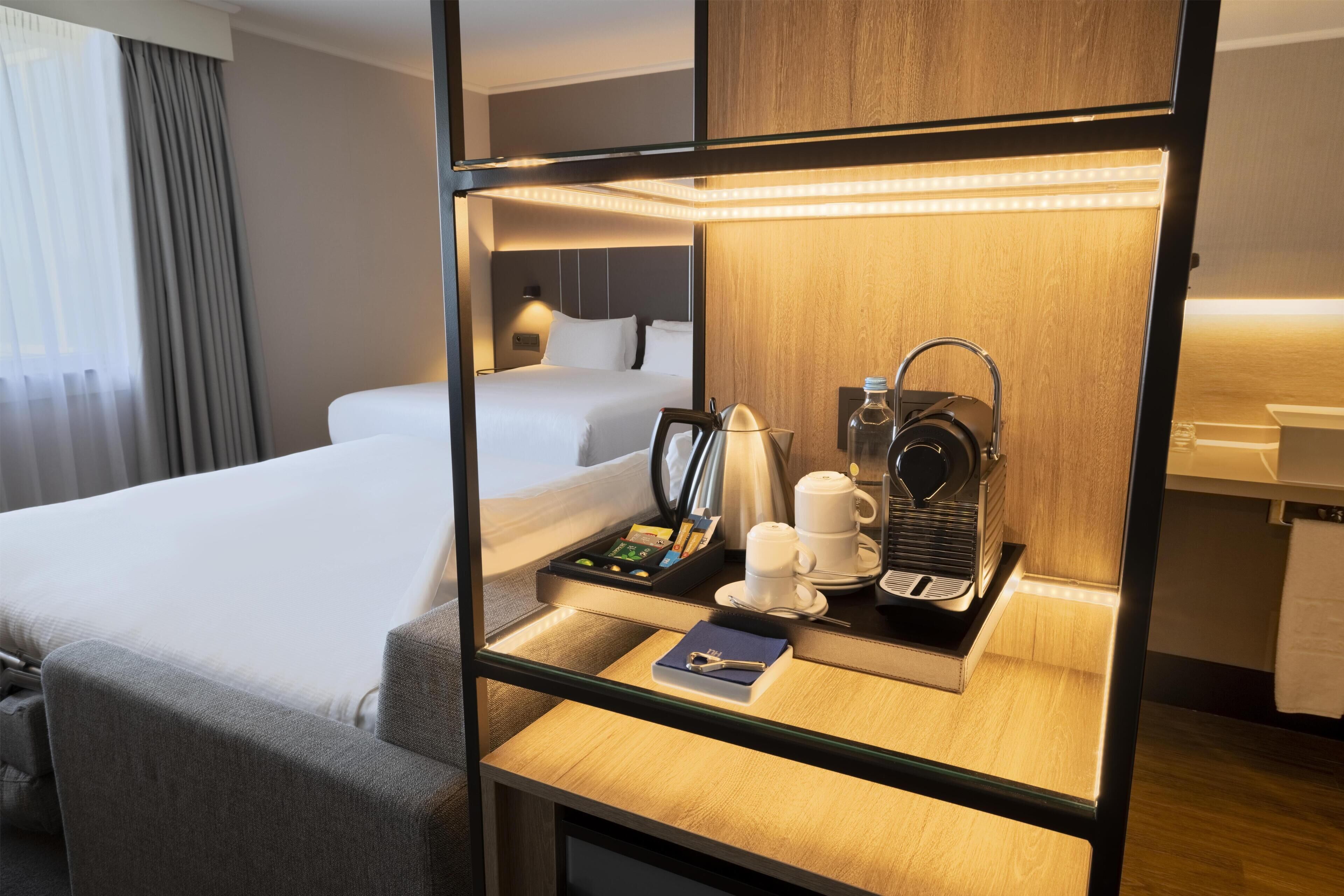 A hotel room with a bed, a bedside table with a kettle, cups, and a tray of snacks.