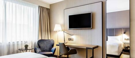 Ett hotellrum med en säng, ett skrivbord, en stol, en tv och en spegel.