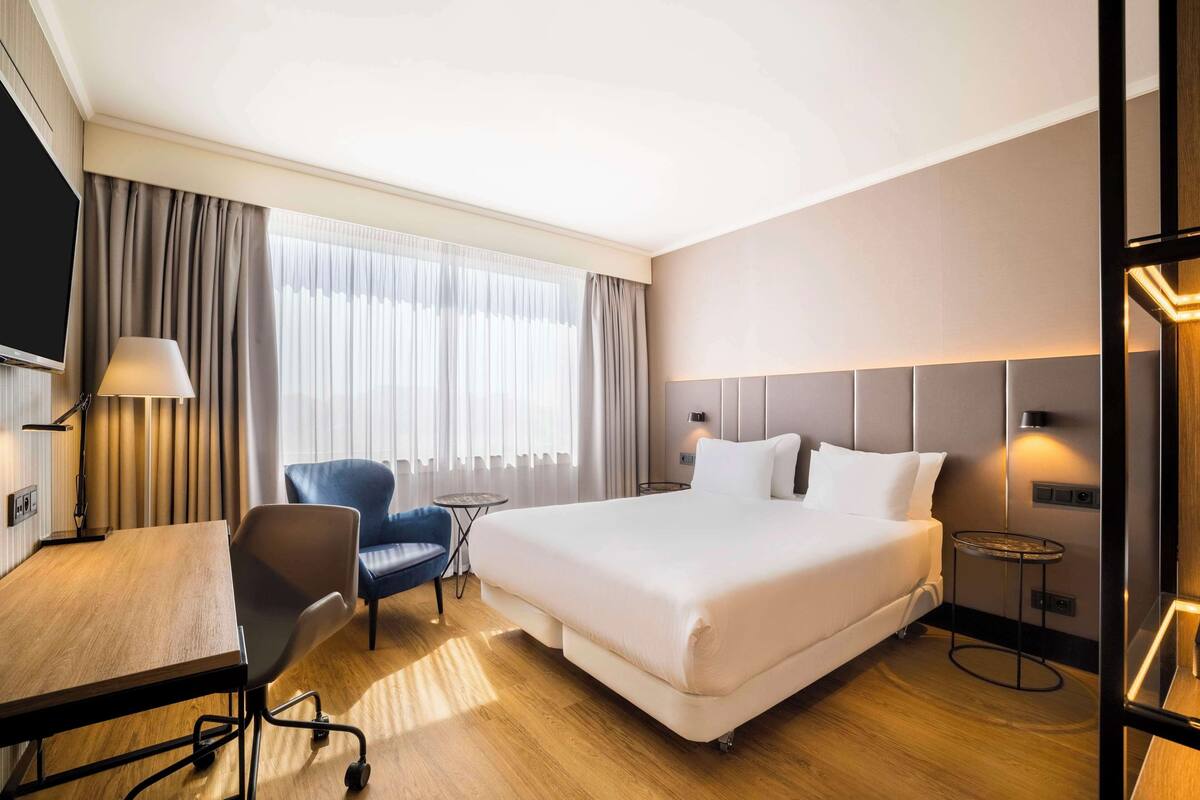 A hotel room with a large bed, a desk with a chair, a small round table, and a TV.