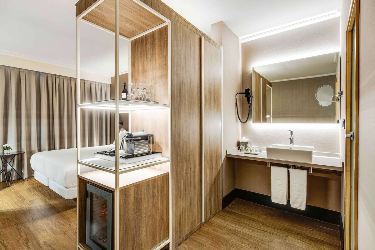 A hotel room with a bed, a desk with a lamp, a vanity with a mirror, and a shower area.