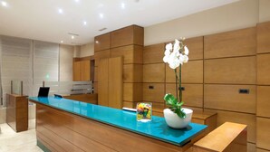 A reception desk with a blue top, wooden paneling on the walls, and a potted plant with white flowers. - NH San Pedro (Marbella)
