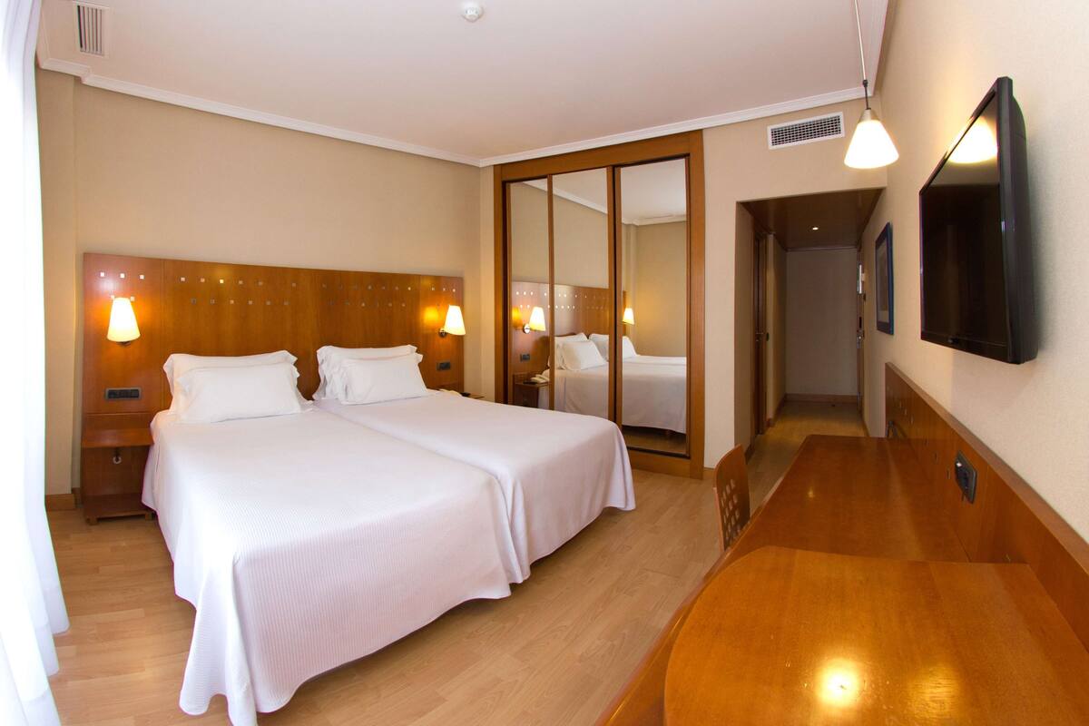 A hotel room with two beds, wooden headboards, bedside lamps, a wooden dresser, and a large mirror.