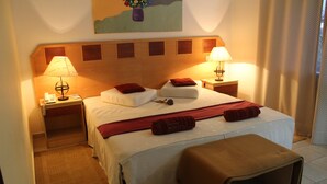 A hotel room with a large bed, two bedside tables with lamps, a headboard with a wooden panel design, a painting on the wall, and a window with curtains. - Hotel Quinta das Pratas (Cartaxo)