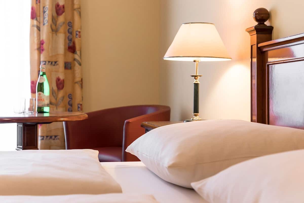 A hotel room with a bed, a chair, a table with a bottle and glasses, and a lamp.