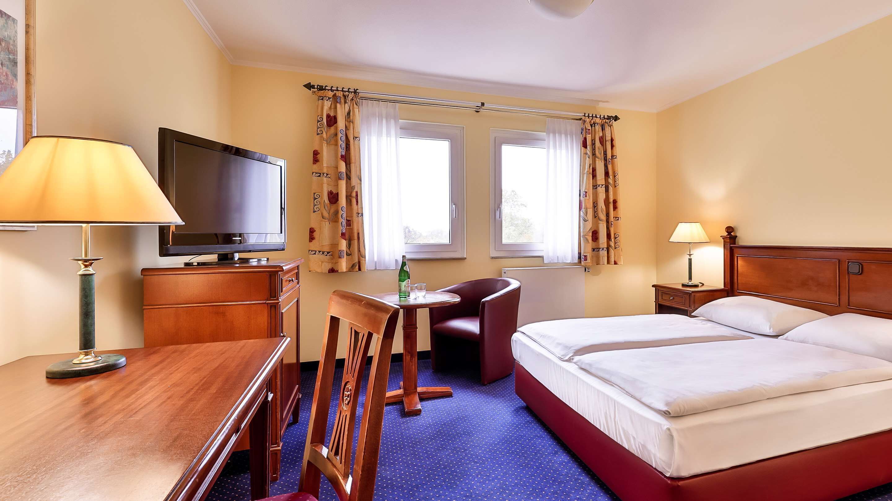A hotel room with a bed, a desk with a television, a chair, and a table.