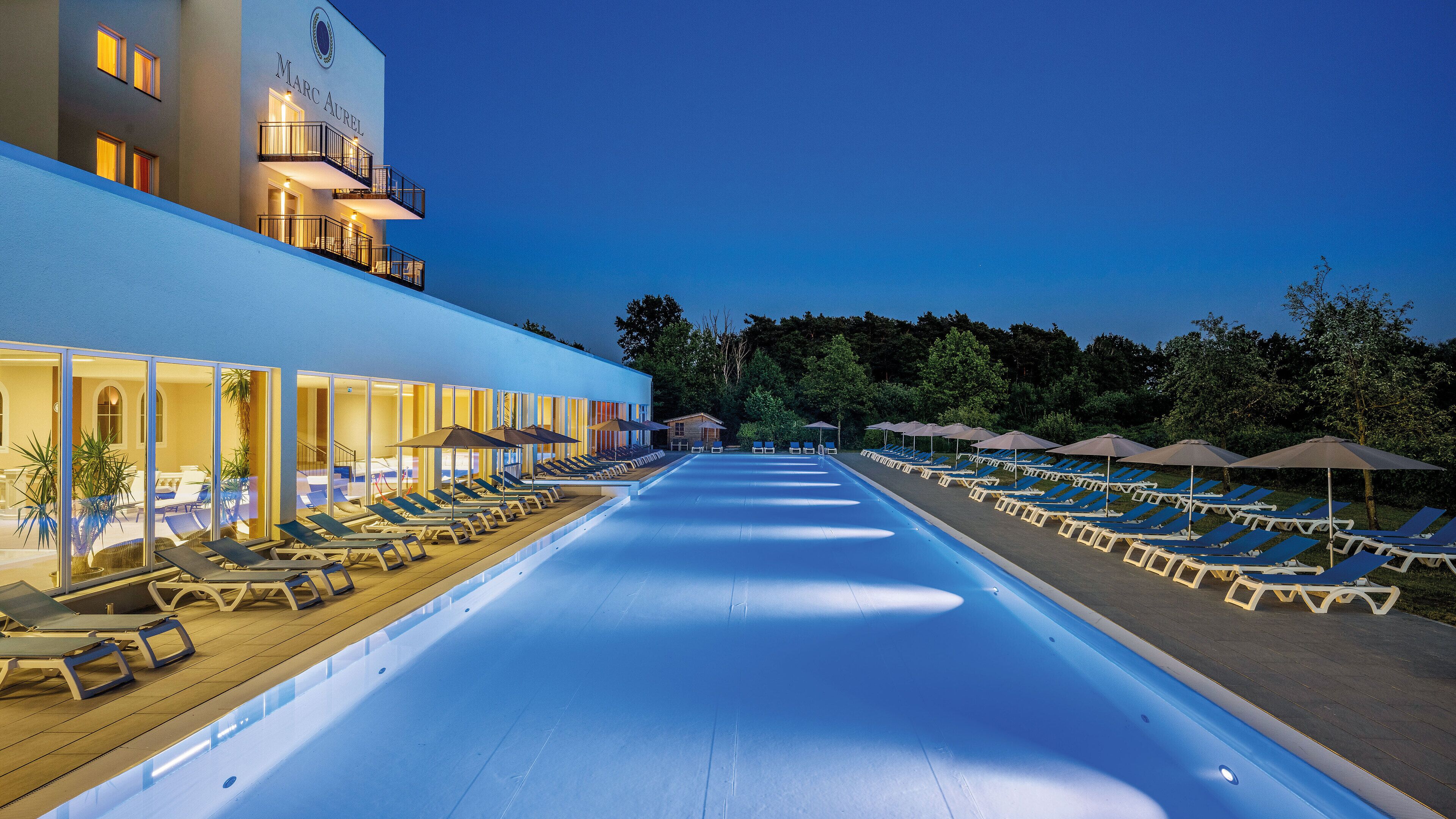 Une piscine avec des chaises longues et des parasols, un bâtiment doté de balcons et un ciel dégagé.