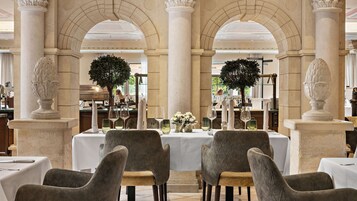 Un restaurante con columnas de mármol, mesas dispuestas con manteles blancos y jarrones decorativos.