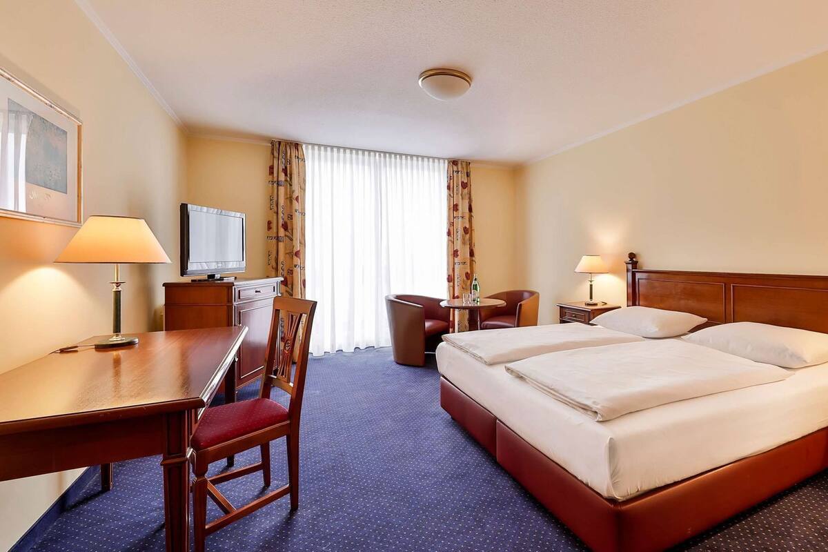 A hotel room with a bed, desk, chair, TV, and a window with curtains.