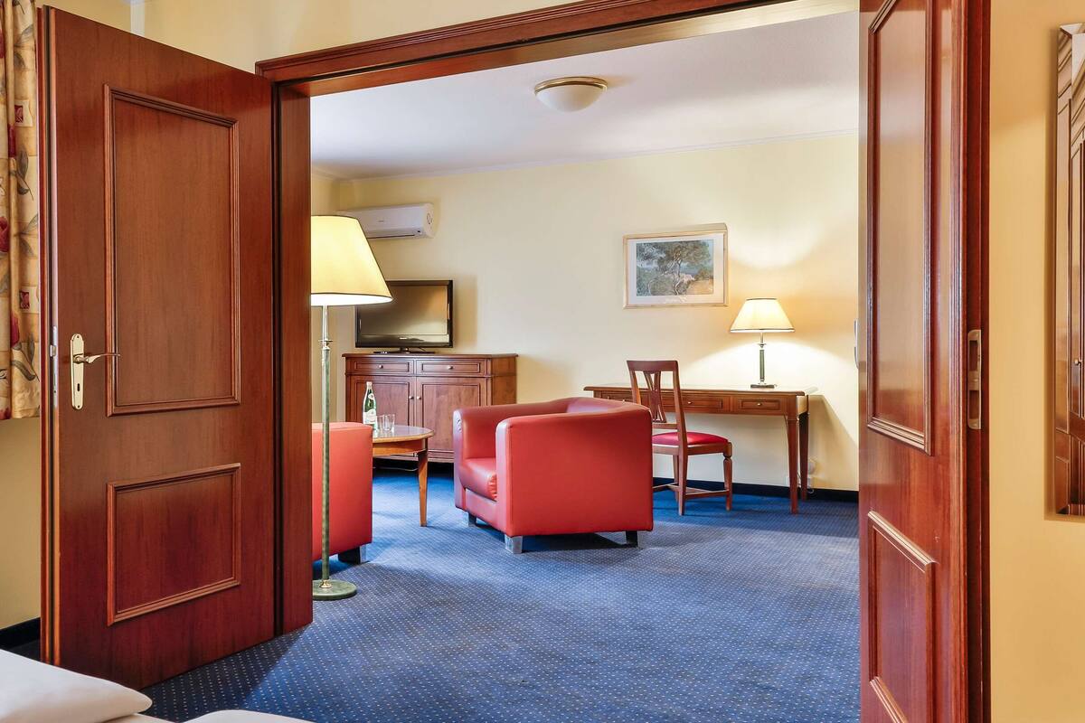 A hotel room with a wooden door, a bed, a desk, a chair, and a television.