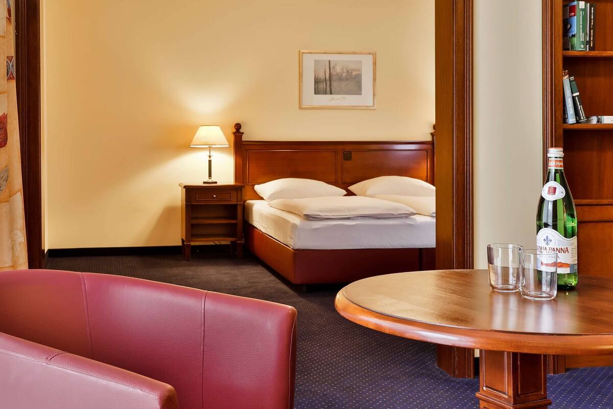 A hotel room with a bed, a nightstand with a lamp, a table with two glasses, and a bottle of water.