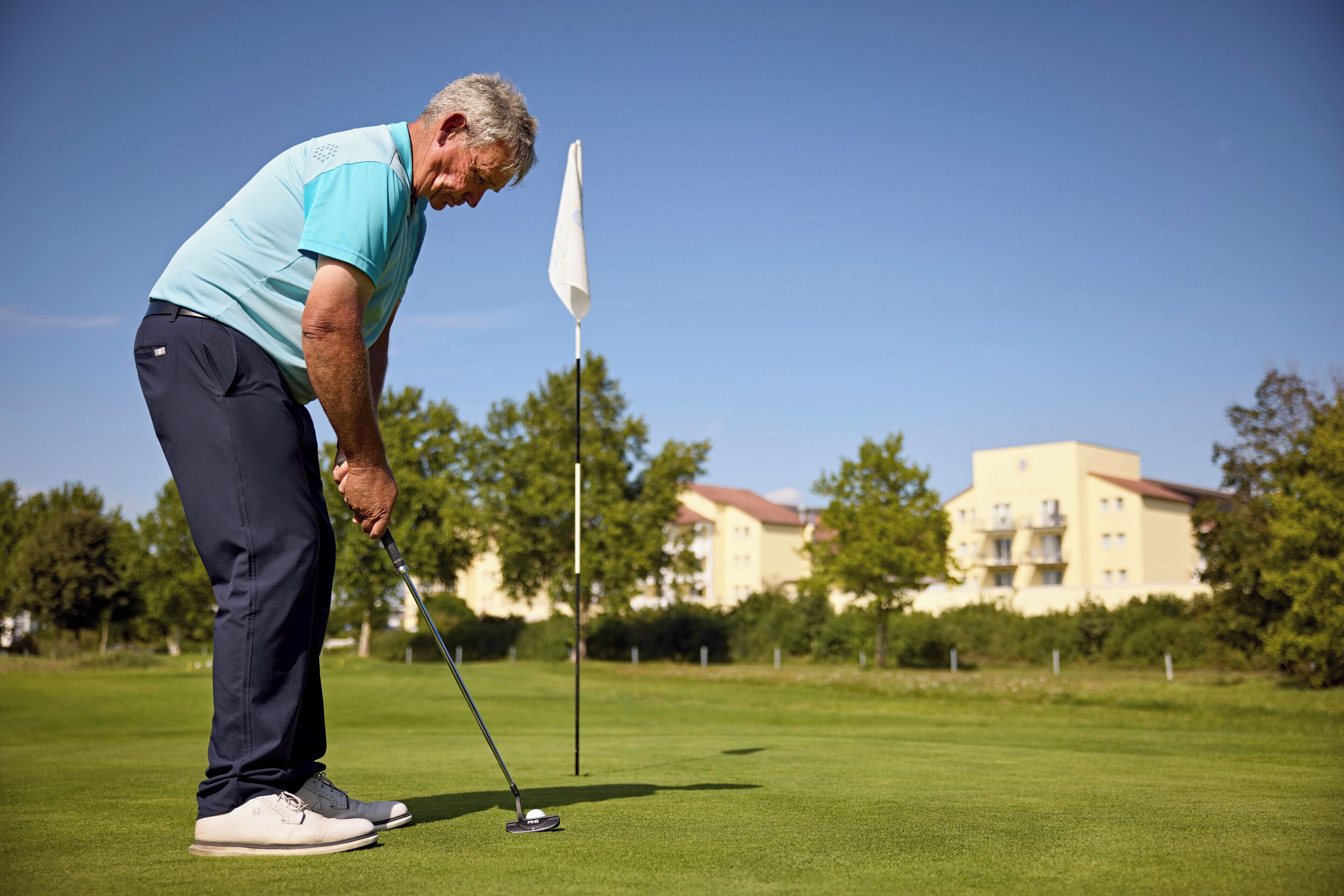 A man in a light blue shirt and dark pants preparing to put a golf ball on a green.